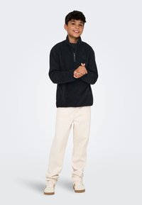 Svart fleece-pullover med høy krage og halv glidelås, paret med kremfargede bukser og lyse sneakers. Glatt tekstur og uformell stil.