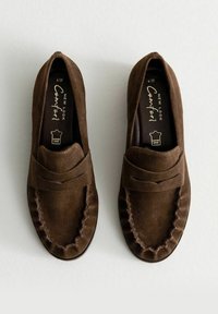 Un paio di mocassini in suede marrone con dettagli cuciti e etichetta in pelle, mostrati dall'alto su uno sfondo chiaro.