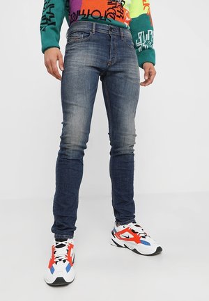 Mann trägt verblasste, eng anliegende blaue Jeans, bunten Grafikpullover und weiße Sneaker mit roten, blauen und schwarzen Akzenten, steht auf einem weißen Boden.