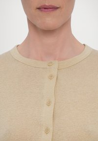 Gros plan d'une personne portant une chemise beige boutonnée avec quatre boutons visibles, montrant l'encolure et le bas du visage.