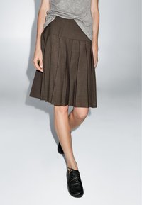 Bruine geplooide rok van textiel, met een hoge taille en een schuin naad. G draagt het met zwarte veterschoenen.