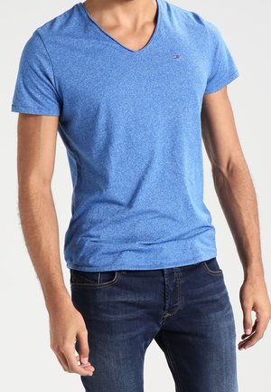 Mies, joka pukeutunut istuvaan siniseen V-kaulaiseen T-paitaan ja tummaan sinisiin farkkuihin, seisoo vaalean yksinkertaisen taustan edessä.