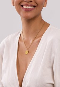 Goldene Halskette mit einem ovalen Anhänger, der ein filigranes, strukturiertes Design aufweist, getragen auf nackter Haut, kombiniert mit einem weißen Leinenoberteil.