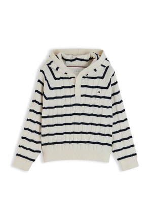 Ragazzo giovane sorridente, indossa un berretto blu navy, un maglione bianco con strisce nere, jeans larghi blu e scarpe da ginnastica bianche, in piedi con le mani in tasca.