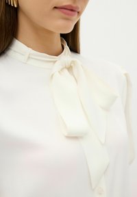 Blusa bianca con un fiocco legato al colletto, caratterizzata da una texture liscia e delicati dettagli sui bottoni, che trasmette un design pulito ed elegante.