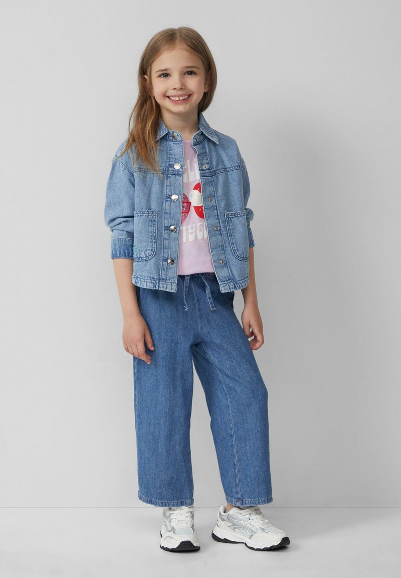 Denimjack in lichtblauw met zilveren knopen, een roze graphic t-shirt eronder, en losse blauwe denim broek in combinatie met witte sneakers.