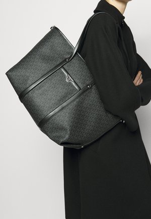 Osoba w czarnym płaszczu niosąca dużą czarną torbę na ramię z wzorem i skórzanymi paskami.