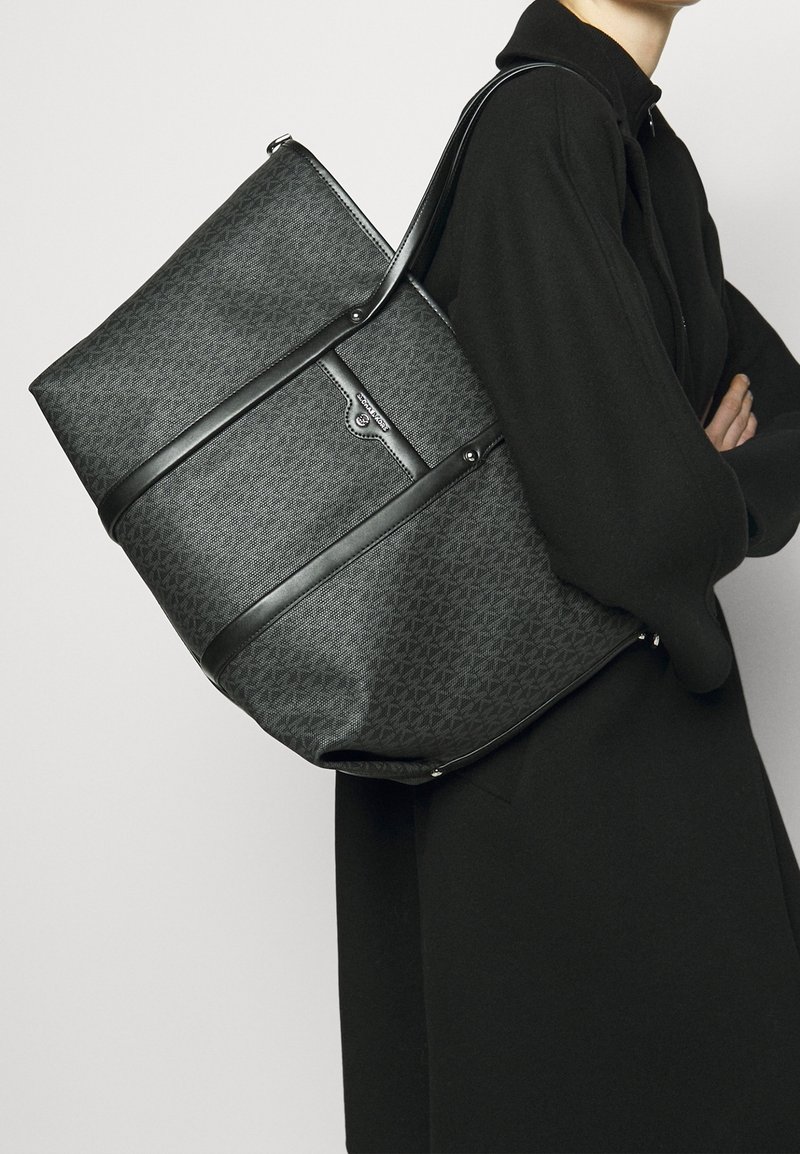 Osoba w czarnym płaszczu niosąca dużą czarną torbę na ramię z wzorem i skórzanymi paskami.