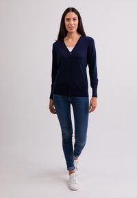 Námořnický svetr, výstřih do V, dlouhé rukávy, zapínání na knoflík, měkká pletená textura, kombinovaný s těsnými modrými džíny a bílými teniskami.