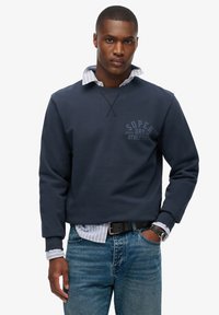 Donkerblauwe sweatshirt met ronde hals en lange mouwen, met een klein grafisch logo. Draagt over een gestreept overhemd met kraag. Spijkerbroek.