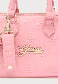 Ροζ συνθετική τσάντα χειρός με ανάγλυφο λογότυπο, επίχρυσα μεταλλικά στοιχεία και αποσπώμενο λουράκι που φέρει ετικέτα. Κλείσιμο με φερμουάρ στην κορυφή.