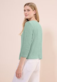 Pull en maille vert clair avec un col rond et des manches trois-quarts, assorti à un pantalon blanc. Aucune motif ou matériel visible.