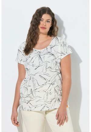 Donna con capelli castani ondulati che indossa una camicia bianca con disegni di uccelli a contorno nero e pantaloni chiari, in piedi contro un muro bianco.