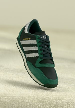 Baskets Adidas vert foncé et noir avec des rayures argentées et des lacets, positionnées au-dessus d'une surface texturée claire.