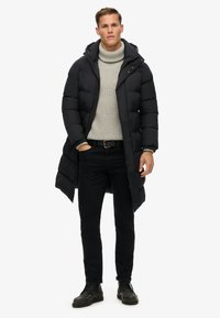 Abrigo tipo puffer negro con capucha, diseño ajustado, tela texturizada, de longitud completa, sobre un cuello de tortuga gris, combinado con jeans negros y zapatos negros.