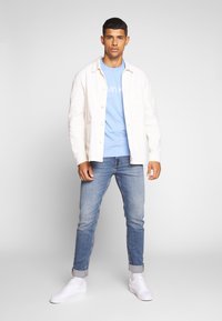 Vyras, dėvintis šviesiai mėlyną Calvin Klein marškinėlį, baltą švarką, sumištas mėlynas džinsus ir baltus sportbačius, stovi prieš paprastą pilką foną.