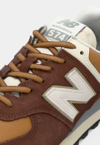 Baskets New Balance 574 marron et beige avec des lacets beige, logo blanc "N" sur le côté et étiquette sur la languette indiquant "New Balance 574."