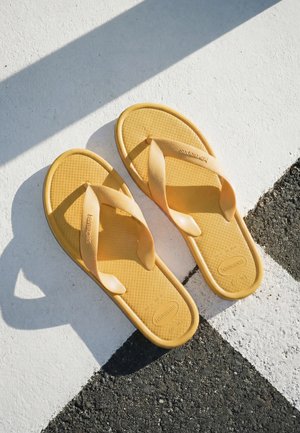 Żółte klapki Havaianas spoczywające na białej i czarnej malowanej nawierzchni asfaltowej, na której światło słoneczne rzuca cienie.