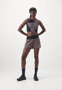 Sportlicher Crop-Top aus lila und schwarzem Stoff mit figurnaher Passform, kombiniert mit lockeren braunen Shorts. Eine schwarze Mütze und Schuhe runden das Outfit ab.