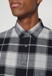 Černá a šedá kostkovaná košile na knoflíky s klasickým límečkem, vyrobená z texturované tkaniny, s dvěma šedými knoflíky u krku.