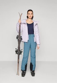 Ženska v sivem jakni, temno modrem vrhu in modrih smučarskih hlačah drži smuči in nosi črne smučarske čevlje na enobarvni podlagi.