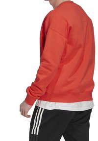 Man med en röd långärmad sweatshirt över en vit skjorta, och svarta byxor med tre vita ränder på sidan.