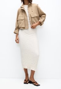 Veste beige coupée recadrée avec deux poches avant sur une robe midi blanche texturée. Le mannequin porte des sandales à talons blocs noirs avec un motif écaille de tortue.