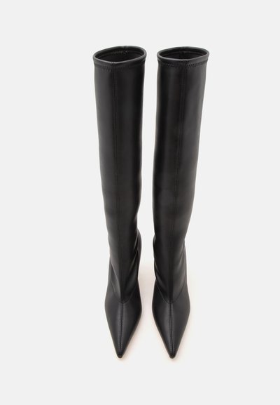 Paire de bottes hautes en cuir noir avec des bouts pointus, montrées debout sur un fond blanc uni.