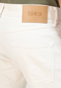 Pantalones de mezclilla blanca con una superficie texturizada, que cuentan con un parche de cuero etiquetado como "EDWIN" y un bolsillo trasero con discretos detalles de costura.