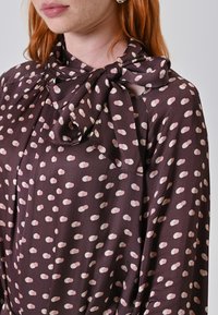 Braune Bluse mit weißen Punkten und einer gebundenen Schleife am Ausschnitt. Der Stoff wirkt glatt und leicht, mit rosa elliptischen Mustern über die gesamte Fläche verteilt.