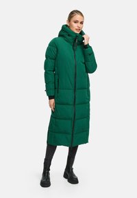 Cappotto imbottito verde con colletto alto, zip frontale e dettagli neri. Ha un design lungo e dritto e presenta cuciture verticali.
