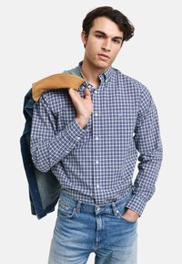 Camisa de cuadros azul y blanca de botones con cuello, combinada con jeans de mezclilla azul claro, con una chaqueta de mezclilla colgada de un hombro.