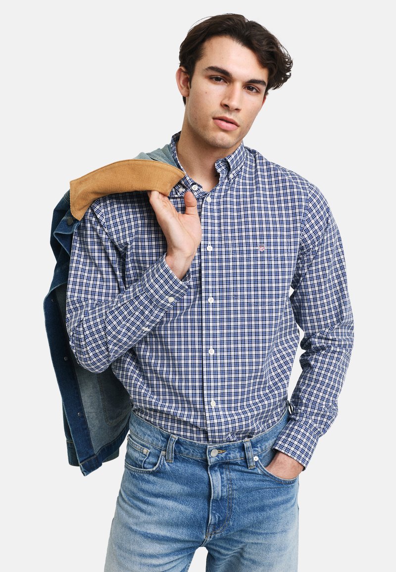 Camisa de cuadros azul y blanca de botones con cuello, combinada con jeans de mezclilla azul claro, con una chaqueta de mezclilla colgada de un hombro.