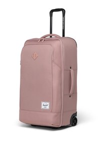 Jemně růžový kufr na kolečkách s texturovanou tkaninou, dvojitými horními uchy, skládanou černou rukojetí a bílou značkovou etiketou na přední straně.