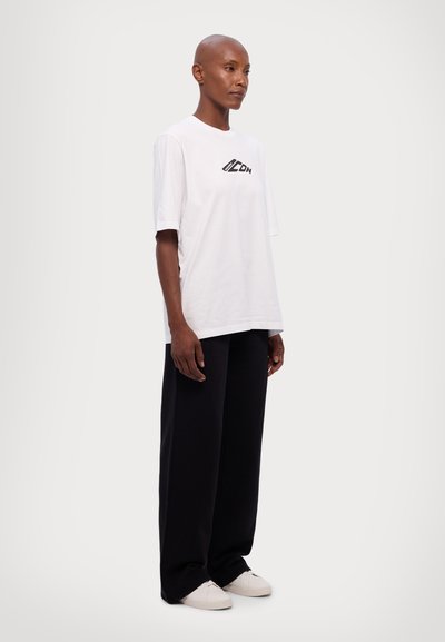 Weißes Baumwoll-T-Shirt mit einem schwarzen Grafik-Logo auf der Brust, kombiniert mit schwarzen weiten Hosen und weißen Sneakers. Einfarbigem Hintergrund.