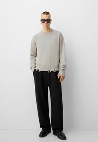 Hellgrauer Strickpullover mit fransigem Saum und Ärmeln, kombiniert mit lockeren schwarzen Hosen und schwarzen Schuhen. Das Modell trägt außerdem eine dunkle Sonnenbrille.