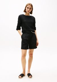 Svart strikket genser med lange ermer, kombinert med skreddersydde svarte shorts med sidelommer og oppbrettede kanter. Svarte sandaler med stropper.