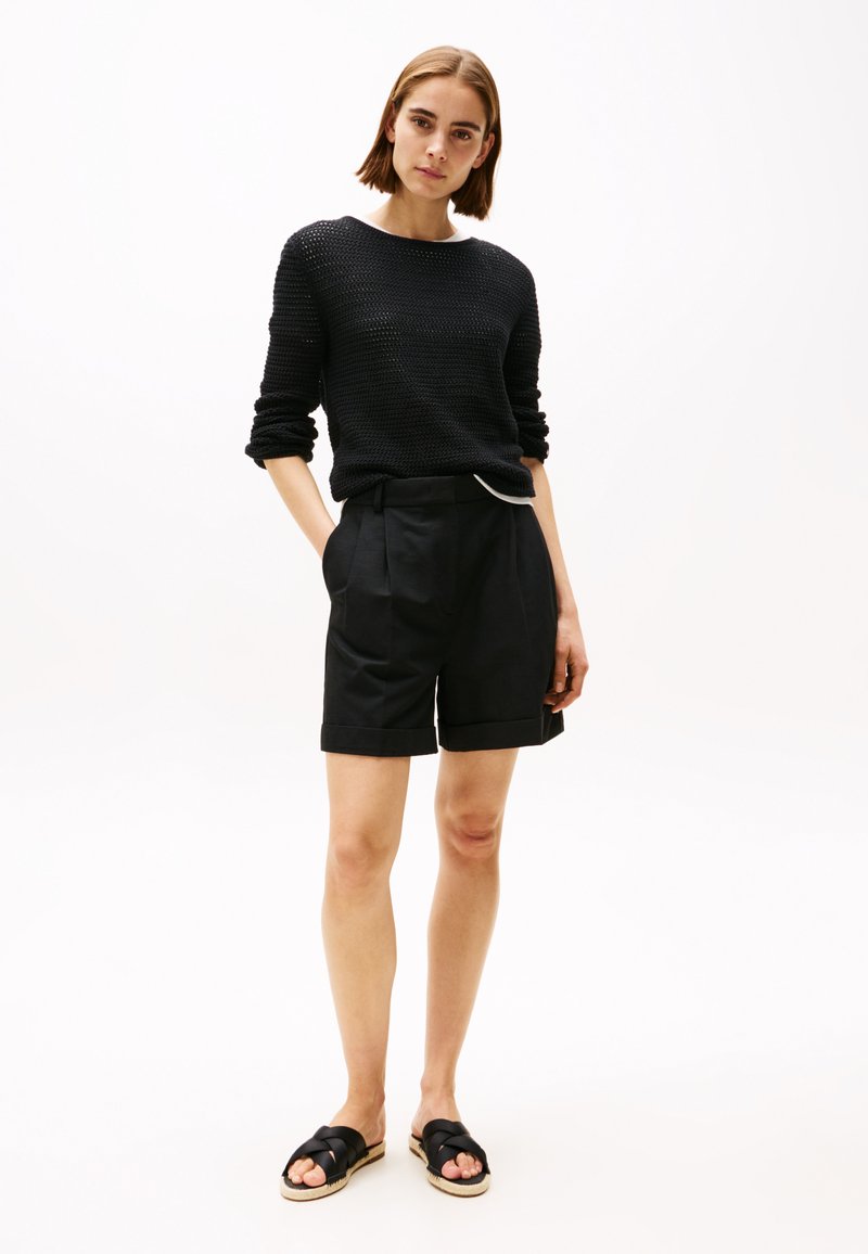 Suéter negro de punto con mangas largas, combinado con pantalones cortos negros ajustados que tienen bolsillos laterales y un dobladillo doblado. Sandalias negras con tiras.