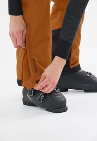 Bruine waterdichte broek met een verstelbare rits bij de enkel, gecombineerd met zwarte skischoenen met dubbele gesp.