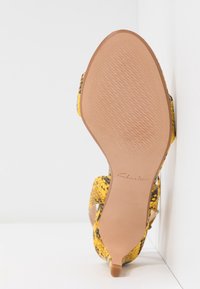 Vue de la semelle d'une sandale à motif peau de serpent jaune et noir, avec un petit talon, une semelle texturée et le logo Clarks sur la semelle.