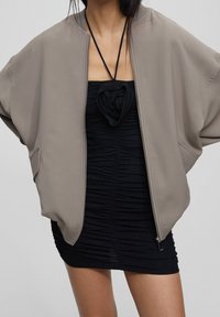 Chaqueta bomber beige de corte holgado en tejido suave, con dos bolsillos frontales y cremallera. Llevada sobre un vestido negro ajustado con fruncidos y un detalle floral.