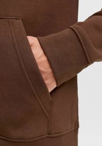 Matière de sweat-shirt marron avec une coupe décontractée. Possède une poche kangourou et des manches côtelées, montrant une main à l'intérieur de la poche.