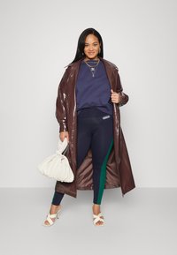 Glansig brun lång jacka, marinblå sweatshirt, marinblå leggings med grön rand, vit handväska och vita klacksandaler.