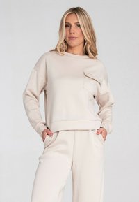 Jasno beżowy sweatshirt wykonany z gładkiego materiału. Posiada przednią kieszeń, okrągły dekolt oraz ściągacze przy mankietach. W zestawie ze spodniami w tym samym kolorze.