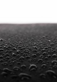 Surface noire et texturée avec de nombreuses gouttes d'eau de tailles variées, réfléchissant la lumière. Le matériau lisse semble résistant à l'eau.
