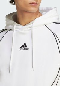 Witte hoodie van zachte stof met zwarte accenten. Voorzien van trekkoorden en geborduurd Adidas-logo op de borst. Gecontourde ontwerp lijnen.