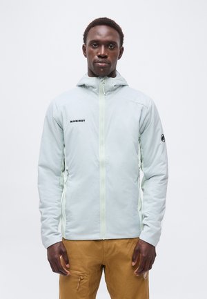 Mann trägt eine hellgraue Mammut Zip-Up-Jacke mit Kapuze und braune Hose, steht vor einem weißen einfarbigen Hintergrund.