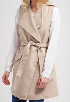 Femme portant un gilet sans manches beige ceinturé sur une chemise blanche à manches longues et un jean bleu devant un fond uni.