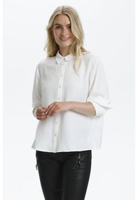 Chemise blanche à boutons avec un col évasé, en tissu doux et manches retroussées. Les détails notables incluent une coupe décontractée et un léger éclat.