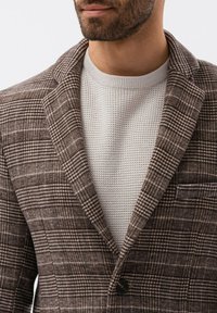 Ruskea ruudullinen bleiseri, jonka alla on tekstuuri-neuleinen kermaun suruvaihetta. Bleiserissä on notched-kaulus ja yksi musta nappikiinnitys.